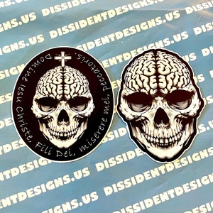 May include: Two stickers featuring skull designs. One is a skull with a brain and a cross, surrounded by text. The other is a skull with a brain. Both are black and white with intricate details, set against a blue background.