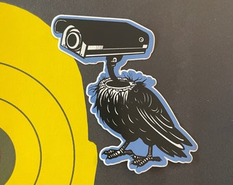 May include: A black and white sticker of a bird with a security camera on its head. The bird is perched on a blue background.