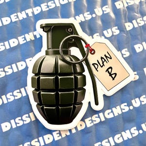 May include: A green and black grenade with a white tag attached to the pin. The tag says "PLAN B" in black lettering.