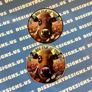May include: Two round stickers featuring a cow wearing a brown mask, surrounded by flowers. The text "NOT A BIG FAN OF THE FDA" is in a yellow arc around the cow. The background is a light blue with white text.