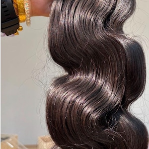 Puede incluir: Un paquete de extensiones de cabello castaño oscuro con textura ondulada. El cabello está sujeto en la parte superior con una banda de tejido negra. Los mechones muestran un brillo natural y están peinados en ondas sueltas y voluminosas.