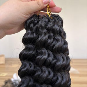 Może przedstawiać: Pęczek czarnych, falowanych przedłużeń włosów, spiętych złotą nicią. Włosy mają naturalny, obszerny wygląd z wyraźnymi lokami. Przedłużenia mają na celu dodanie długości i objętości.