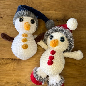 Könnte beinhalten: Zwei gehäkelte Schneemann-Ornamente, eines mit einem blau-grauen Hut und das andere mit einem rot-weißen Hut mit Kunstfellbesatz.