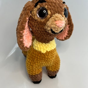 Op de afbeelding: Een bruin gehaakt knuffeldier met grote zwarte ogen en een roze neus. Het dier heeft een gele kraag en staat op een wit oppervlak.