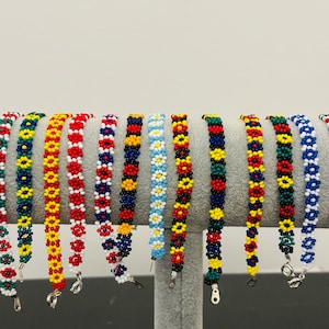 Könnte beinhalten: Eine Sammlung von 14 bunten Perlenarmbändern mit Blumendesigns. Die Armbänder sind aus roten, gelben, blauen, grünen, weißen und schwarzen Perlen gefertigt.