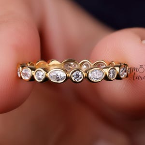 Könnte beinhalten: Ein goldener Ring mit einer Reihe ovaler und runder klarer Edelsteine. Der Ring wird zwischen zwei Fingern gehalten. Der Ring ist im Bandstil gehalten, wobei die Edelsteine eng beieinander liegen. Der Ring hat den Text "Diamond Aura" an der Seite.