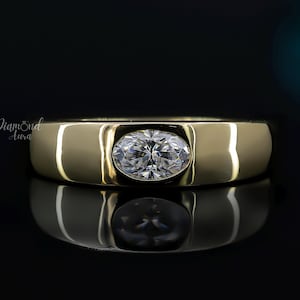 Puede incluir: Anillo de banda dorado con una gema ovalada y transparente en el centro. El anillo se muestra sobre una superficie negra reflectante, con el nombre de la marca "Diamond Aura" visible en el fondo.