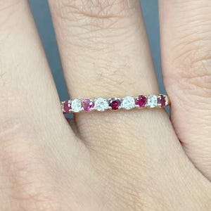 Puede incluir: Un anillo de oro con rubíes y diamantes redondos alternados. Las piedras preciosas están engastadas muy juntas, creando una banda continua de color y brillo. El anillo se muestra en un dedo.