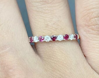 May include: A gold ring featuring alternating round-cut rubies and diamonds. The gemstones are set closely together, creating a continuous band of color and sparkle. The ring is shown on a finger.