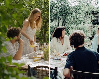 May include: A group of people gathered around a wooden outdoor dining table set with white linens, ceramic plates, bowls of fresh berries, and glassware. The scene features individuals in casual clothing enjoying a meal in a lush garden setting.