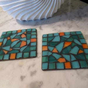 May include: Two square mosaic coasters with a teal, blue, and orange geometric pattern. The coasters are made of small, irregular pieces of glass.