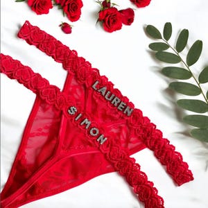 May include: Red lace thong underwear with the names "SIMON" and "LAUREN" spelled out in silver letters. The underwear is decorated with a floral lace pattern. Red roses and greenery are arranged around the underwear on a white surface.