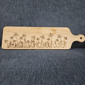 Könnte beinhalten: Rechteckiges Holzschneidebrett mit Griff und Loch zum Aufhängen. Das Brett hat ein eingraviertes Design aus Pilzen und Pflanzen entlang der Oberkante. Das Holz hat eine helle, natürliche Oberfläche.