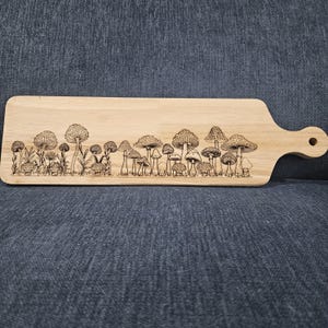 Könnte beinhalten: Ein Holzschneidebrett mit Griff, das ein lasergraviertes Design verschiedener Pilze zeigt. Das Brett ist rechteckig mit abgerundeten Ecken und einer natürlichen Holzoberfläche. Die Pilzillustration erstreckt sich über die gesamte Länge des Brettes.