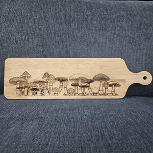 Könnte beinhalten: Ein Holzservierbrett mit Griff, das ein lasergraviertes Design verschiedener Pilze aufweist. Das Brett ist rechteckig mit abgerundeten Kanten und einer natürlichen Holzoberfläche. Die Pilzillustration umfasst detaillierte Kappen und Stiele.