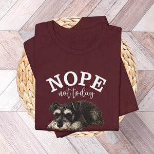 May include: A maroon t-shirt with the words "NOPE not today" in white lettering above a graphic of a Schnauzer dog. The dog is black, gray, and white, lying down.