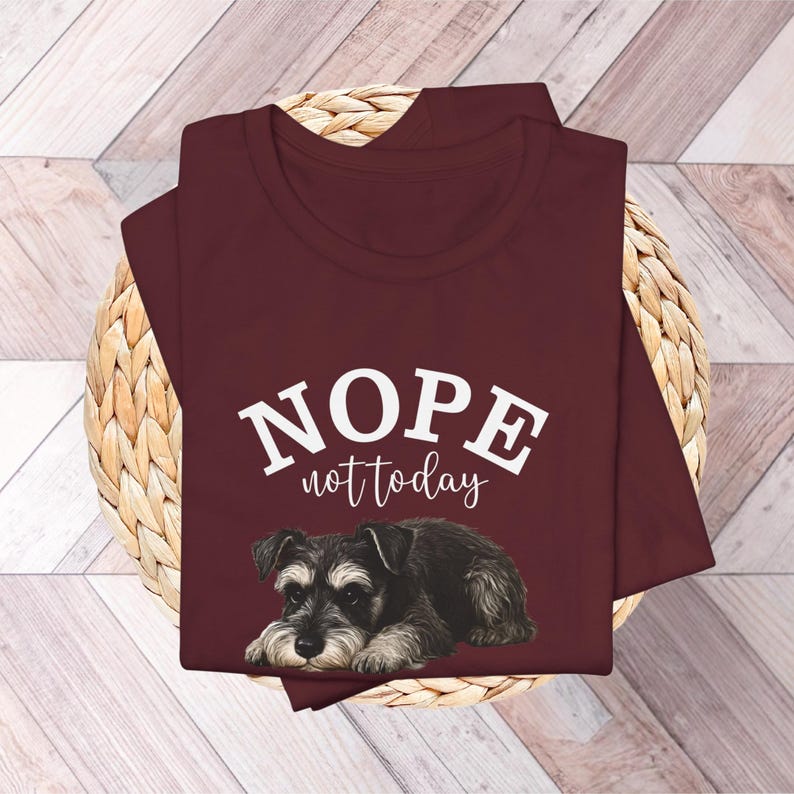 May include: A maroon t-shirt with the words "NOPE not today" in white lettering above a graphic of a Schnauzer dog. The dog is black, gray, and white, lying down.