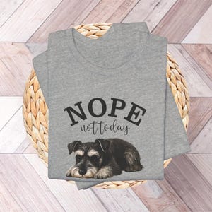 May include: A heather gray V-neck t-shirt featuring the text "NOPE not today" above a detailed illustration of a Schnauzer puppy. The dog is black, gray, and brown, with a relaxed pose. The shirt is folded on a woven basket.
