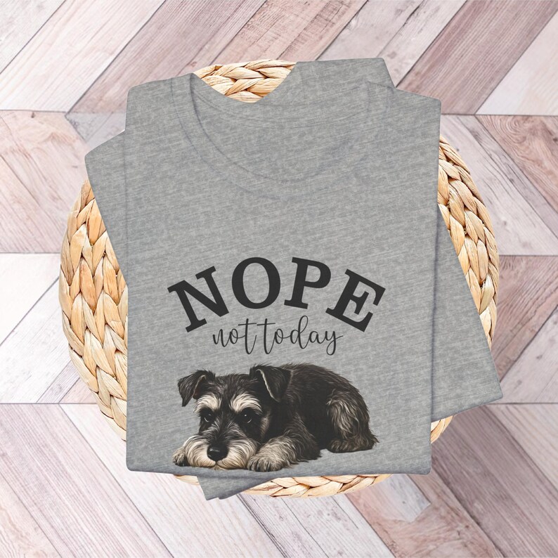 May include: A heather gray V-neck t-shirt featuring the text "NOPE not today" above a detailed illustration of a Schnauzer puppy. The dog is black, gray, and brown, with a relaxed pose. The shirt is folded on a woven basket.