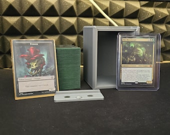 Könnte beinhalten: Eine graue Box mit einem durchsichtigen Plastiketui, in dem sich eine Spielkarte mit dem Text "The Mycosynth" und einer grün-schwarzen Illustration befindet. Neben der Box liegt eine zweite Spielkarte mit dem Text "Fungus" und einer rot-schwarzen Illustration. Hinter der Fungus-Karte befindet sich ein grünes Kartenspiel. Vor der Box liegt ein grauer Plastikteiler mit zwei Löchern.
