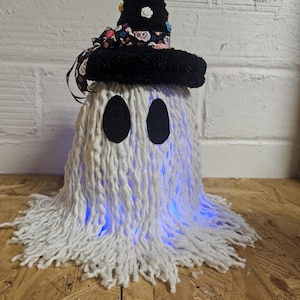 May include: A handmade Halloween decoration shaped like a ghost. The ghost is made of white yarn with black felt eyes and a black witch hat decorated with flowers and ribbon. The ghost glows with blue light.