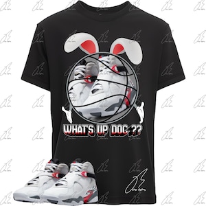 May include: Black t-shirt featuring a basketball with a pair of white and red sneakers inside, bunny ears, and cartoon hands. The text "WHAT'S UP DOC??" is printed below the basketball. A pair of white and red sneakers are at the bottom.