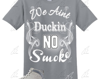 May include: A gray t-shirt with the text "We Ain't Duckin No Smoke" in white lettering. The text is surrounded by a white smoke design. A pair of gray and white sneakers are shown below the t-shirt.