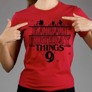 May include: Red t-shirt with black text and graphics. The shirt features the words "KAIRAH BIRTHDAY THINGS 9" in a bold font, with silhouettes of cyclists above the text. The shirt is a solid red color.