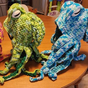 May include: Crocheted octopus hats in shades of green, blue, and pink. Each hat features large, round eyes and multiple dangling tentacles. The hats are displayed on a wooden table, showcasing the handmade craftsmanship.