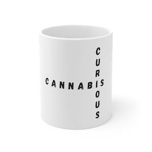May include: White ceramic mug with the words "CANNABIS" and "CURIOUS" in black, arranged in a cross shape. The mug is cylindrical and appears to be empty.