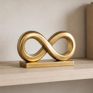Op de afbeelding: Een goudkleurige oneindigheidssymboolsculptuur op een rechthoekige basis, tentoongesteld op een houten plank. De plank bevat ook een witte vaas met groen, een klein rond object, boeken en een glazen kom.