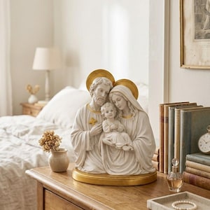 May include: A detailed statue of the Holy Family, featuring Joseph, Mary, and the infant Jesus. The figures are depicted in flowing robes with gold halos. The statue is set on a gold base and placed on a wooden surface.