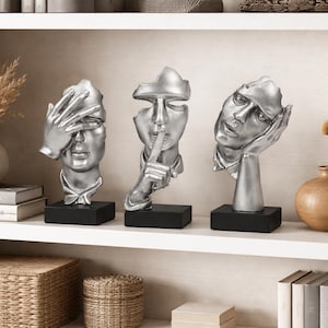 May include: Three silver-colored abstract face sculptures on black bases, each depicting a different gesture: covering eyes, finger to lips, and hand to cheek. The sculptures are displayed on a white shelf with decorative vases and books.