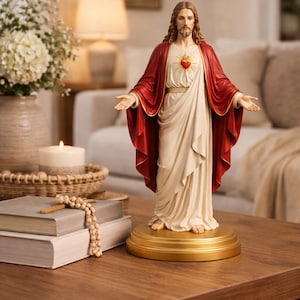 Könnte beinhalten: Religiöse Jesus-Statue mit ausgestreckten Armen, in einem weißen Gewand und einem roten Umhang. Die Figur steht auf einem goldenen Sockel mit einem Herz-Emblem. Auf einem Holztisch platziert.