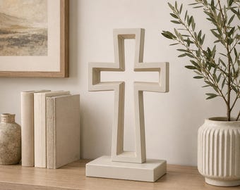 May include: A cream-colored, openwork cross sculpture on a rectangular base. The cross is displayed on a wooden surface, with a framed landscape painting and decorative books in the background. A vase with greenery is also present.