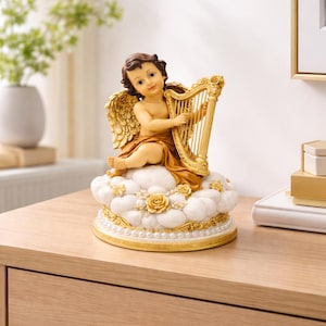 Könnte beinhalten: Dekorative Figur eines Cherubs, der eine goldene Harfe spielt. Der Cherub sitzt auf einer wolkenförmigen Basis, die mit Perlen und Rosen verziert ist. Die Figur hat goldene Flügel und steht auf einer Holzoberfläche.