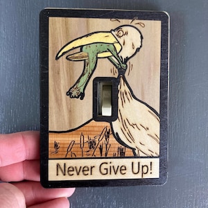 Puede incluir: Cubierta de interruptor de luz de madera con un pájaro sosteniendo una rana, con las palabras "Never Give Up!" debajo. El pájaro y la rana son verdes y amarillos, sobre un fondo claro de vetas de madera con un borde negro.