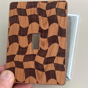 May include: A wooden light switch cover with a checkerboard pattern in shades of brown. The cover has a rectangular opening for the switch and is partially covering a silver metal switch box.