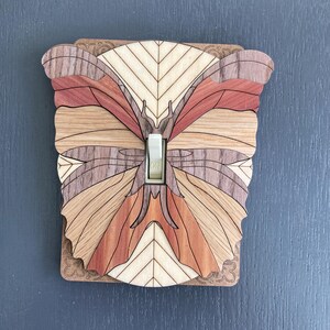 Könnte beinhalten: Eine Holz-Lichtschalterabdeckung mit einem Schmetterlingsdesign. Der Schmetterling besteht aus verschiedenen Holztönen, darunter hellbeige, rotbraun und mittelbraun. Der Schalter ist weiß, vor einem dunkelgrauen Hintergrund.