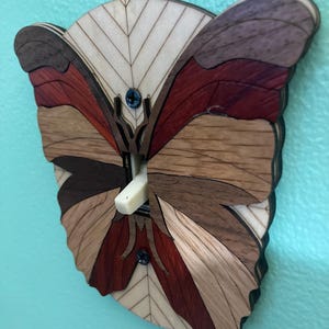 Könnte beinhalten: Ein Lichtschalter-Abdeckung aus Holz mit einem Schmetterlingsdesign. Der Schmetterling besteht aus verschiedenen Holzarten, wodurch ein mehrfarbiges Muster entsteht. Die Flügel des Schmetterlings sind ausgebreitet und der Schalter befindet sich in der Mitte des Schmetterlingskörpers.