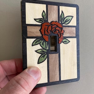 Puede incluir: Cubierta de interruptor de luz de madera con una rosa roja y hojas verdes. La cubierta tiene un diseño de cruz con paneles de madera clara y oscura. Los bordes son negros. La abertura del interruptor es rectangular.