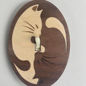 May include: Wooden light switch cover featuring a cat design. The oval-shaped cover has a light wood cat silhouette on a darker wood background. The switch is white and reads "ON".