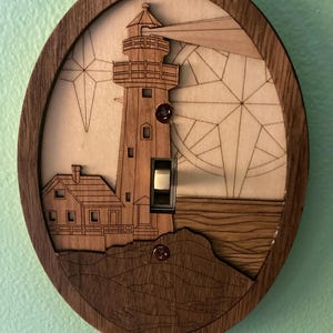 May include: A wooden light switch cover with a laser-cut design of a lighthouse, a house, and a star. The switch cover is oval-shaped and has a dark brown wood finish.