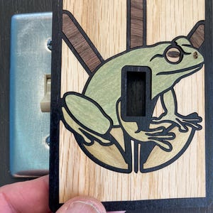 Puede incluir: Cubierta de interruptor de luz de madera con un diseño de rana verde. La rana está colocada sobre un fondo claro de vetas de madera con un borde negro. La cubierta está diseñada para adaptarse a un interruptor de luz estándar.