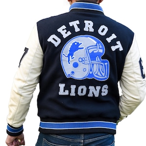 Pode incluir: Uma jaqueta varsity preta com mangas cor de creme, apresentando um grande logotipo bordado de um capacete de futebol americano dos Detroit Lions nas costas. O texto DETROIT LIONS é costurado em branco e cinza, com acabamento canelado listrado em azul e branco na gola, punhos e bainha.