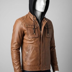 May include: A brown leather jacket with a black hooded sweatshirt underneath. The jacket has two chest pockets and two lower pockets. The jacket is buttoned and zipped up. The mannequin is wearing blue jeans.