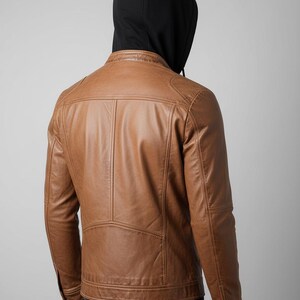 May include: A brown leather jacket with a black hooded sweatshirt underneath. The jacket has a classic design with a stand-up collar, shoulder panels, and vertical stitching details. The jacket is a medium brown color.