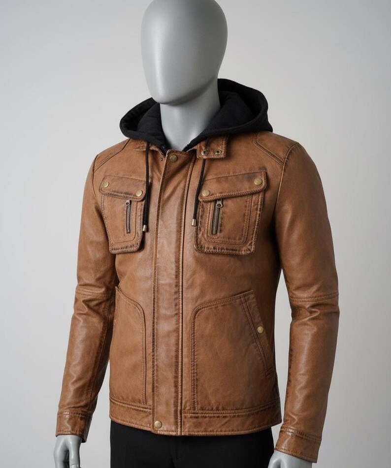 May include: A brown leather jacket with a black hooded lining. The jacket features two chest pockets with zippers, two lower pockets, and a front zipper closure. The jacket is displayed on a mannequin.