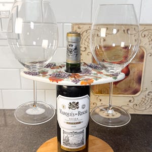 Peut inclure: Un support de bouteille de vin en bois avec deux verres à vin. Le support a un dessus décoratif avec un motif de raisin. Une bouteille de Marqués de Riscal Rioja Reserva 2020 est au centre.