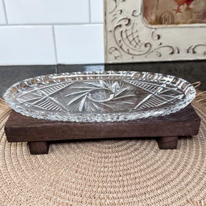 May include: A clear, oval-shaped glass serving dish with a detailed starburst and geometric pattern, resting on a dark brown wooden stand. The stand has four small feet. The background includes a white tiled wall and a framed picture.